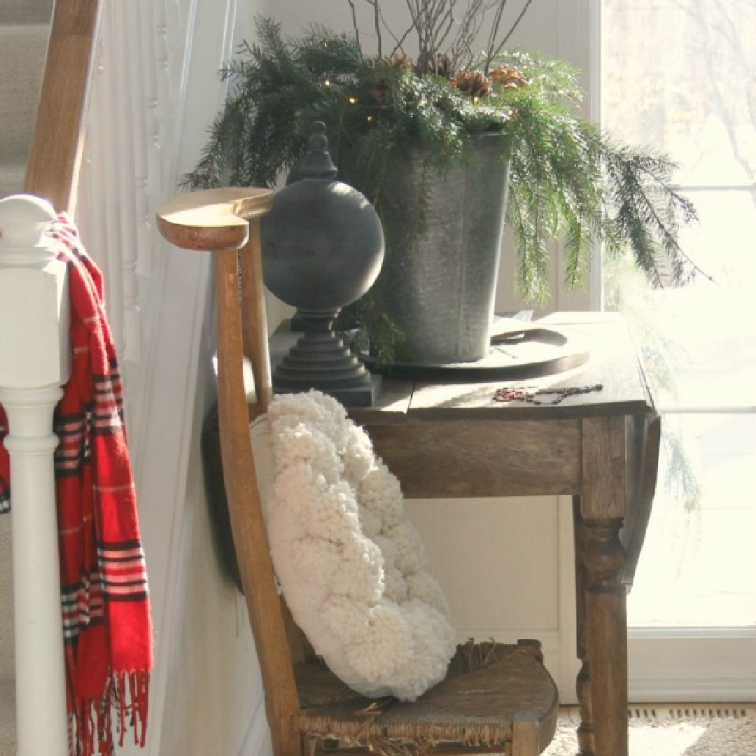 Rustic French country holiday decor in my entry with antique French prayer chair, red scarf, and pom pom wreath - Hello Lovely Studio. #rusticchristmasdecor #christmasentry #hellolovelychristmas