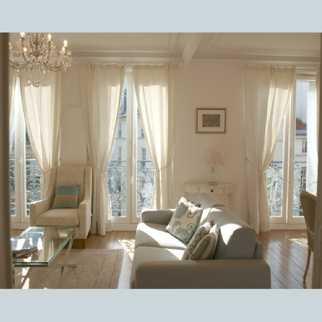 Dappled light softly illuminating a serene Paris apartment living room with hardwood flooring, French doors to balconies, and crystal chandeliers - Hello Lovely Studio.