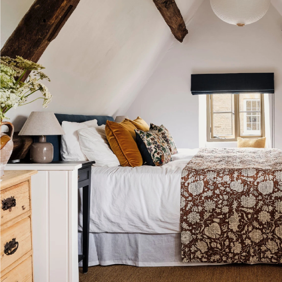 Warm cozy bedroom with rustic ceiling beams in Cotswolds stone cottage. Called Armada Cottage dates to 1489 with reimagined interiors by Beth Dadswell (Imperfect Interiors).